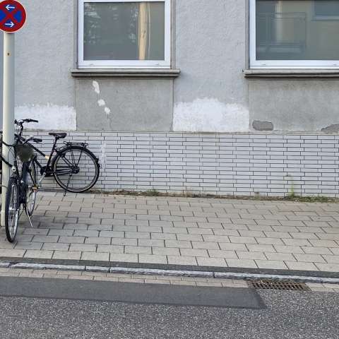 Das Bild zeigt den leeren Platz auf dem Bürgersteig vor den Häusern und neben einem parkenden Auto.